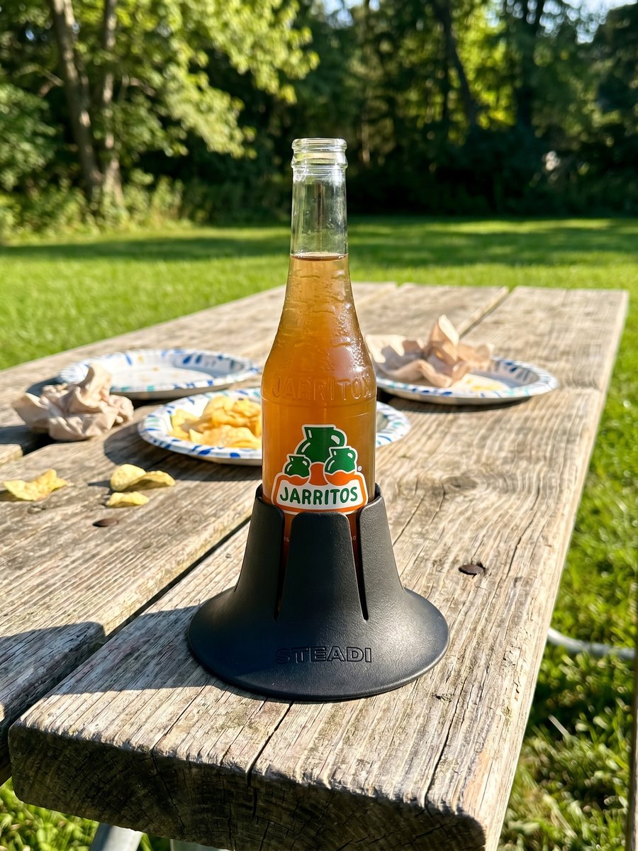 A glass soda bottle standing in a charcoal Steadi on a weathered wooden table outdoors, mown grass and trees behind it.